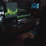A cybersecurity expert in a dimly lit room is typing on a colorful keyboard with multiple screens displaying data.