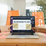 Two professionals strategizing a business plan on a laptop with sticky notes and profit goals.