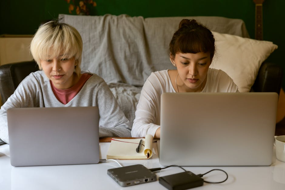 Young ethnic female student surfing internet on laptop while sitting near concentrated friend typing on netbook near hard drives and copybook in apartment