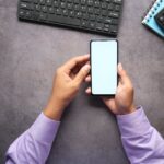 Man in purple shirt holding a smartphone with blank screen at office desk with keyboard, notebook, and calculator.