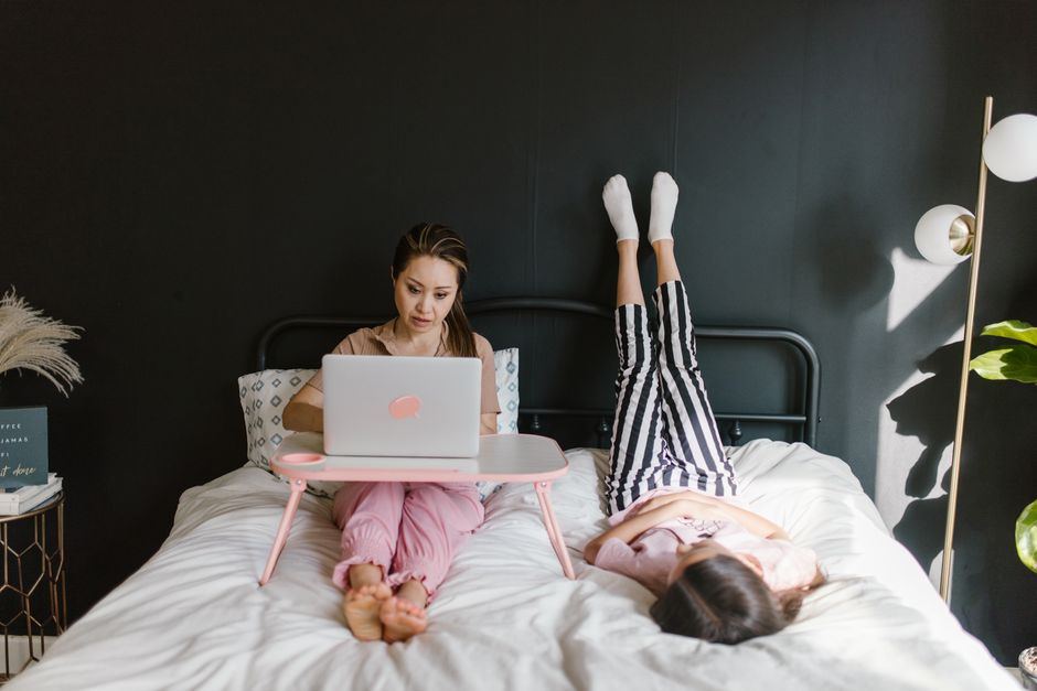 Woman working remotely on laptop in bed, child playing nearby, embracing new work-from-home culture.