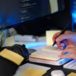 A person planning a software project with handwritten notes and code on a monitor in a modern workspace.